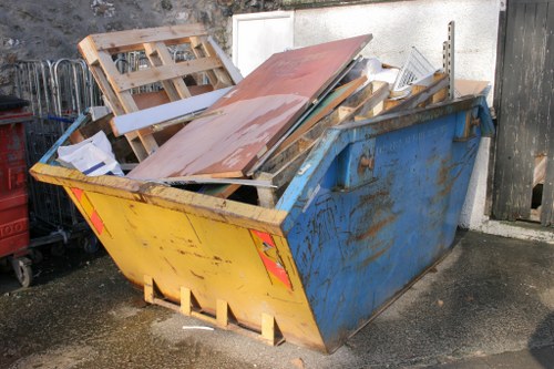 A large blue industrial skip positioned on an asphalt driveway near a concrete wall, filled with various discarded construction and household materials including wooden pallets, metal sheets, and broken furniture. The skip shows rust and signs of use, with some debris spilling slightly onto the ground. To the left, partially visible are additional waste management elements, indicating an active rubbish removal process. The scene is outdoors, surrounded by an urban setting consistent with a central London area, suggested by the context of Marylebone House Clearance. The area appears clean and organized, with natural daylight illuminating the contents of the skip, supported by the presence of nearby fencing and a building facade in the background. This image exemplifies the type of waste collected by Marylebone House Clearance services for rubbish removal and clearance in the Westminster area, including postcode central London.