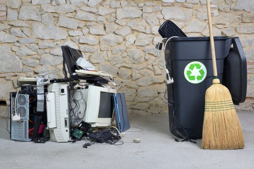 In the foreground, a pile of electronic waste, including outdated computer monitors, keyboards, computer towers, and tangled cables, is situated on a paved surface with some debris scattered around. To the right, a large black rubbish bin featuring a green recycling symbol is positioned against a light-colored stone wall, with a wooden broom leaning against it. The scene indicates a waste collection or clearance area, likely outside a residential or commercial property, associated with rubbish removal services in Marylebone. The overall setting is clean, well-lit, and shows typical materials encountered during a house clearance, emphasizing the disposal of electronic and general waste.