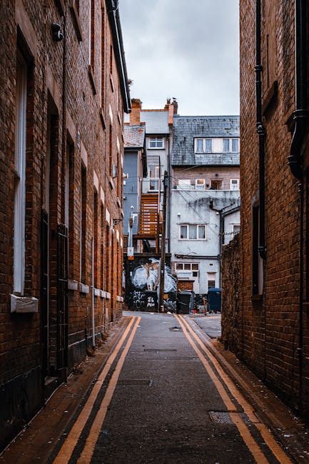 A narrow alleyway situated between two brick buildings, with a tarmac surface marked by double yellow lines along each side. The left building features two white-framed windows and a black drainpipe a